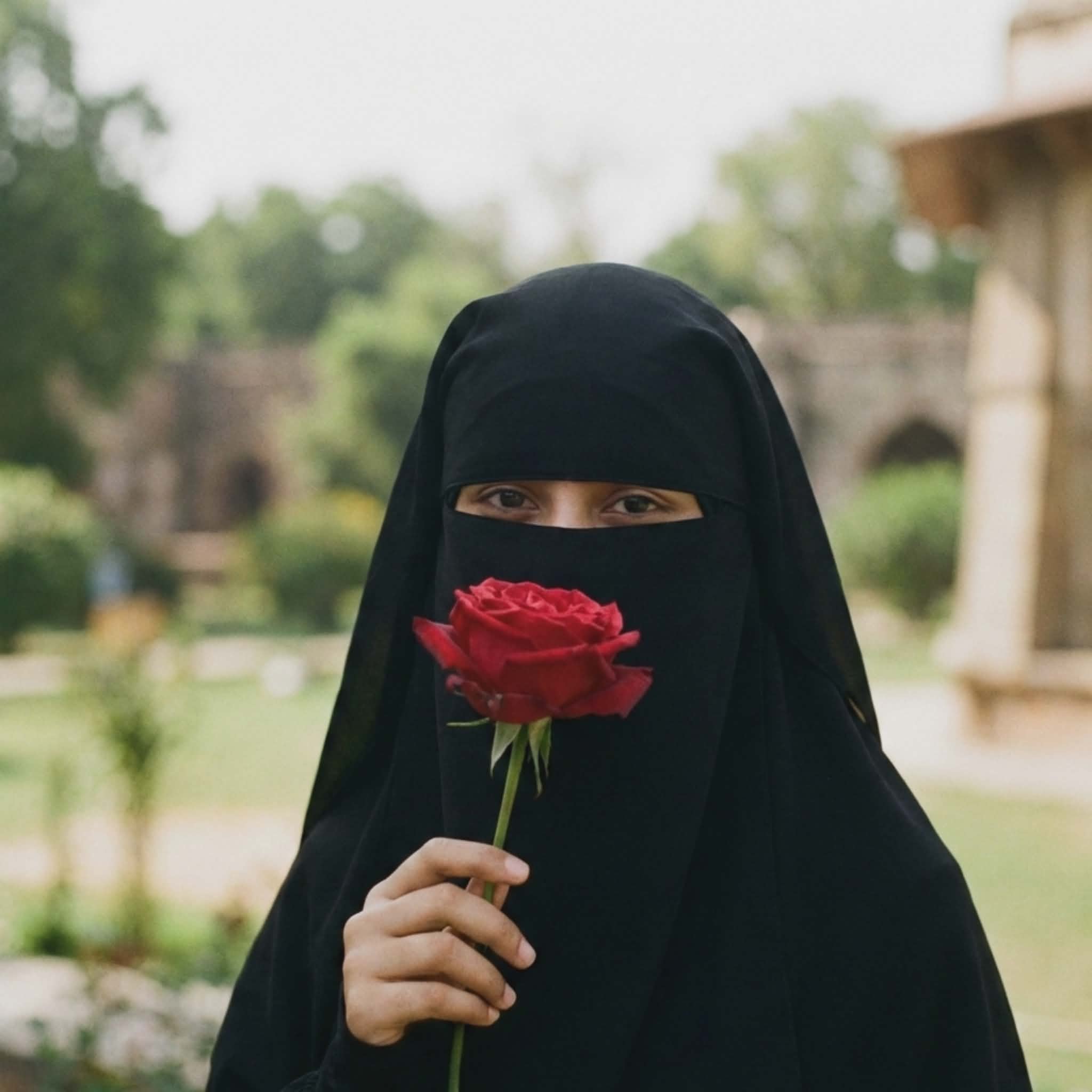 Aesthetic Muslim girl in black niqab holding a red rose in front of her face, beautiful eyes visible, blurred garden background, romantic Islamic DP.