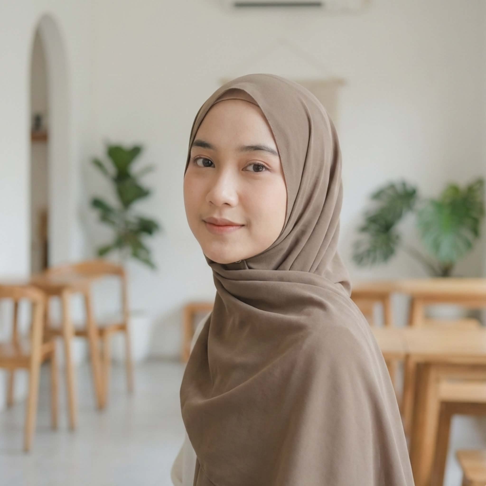 Portrait of a beautiful Muslim girl wearing a dark beige/brown hijab, smiling gently, blurred cafe interior background, aesthetic modest profile picture.