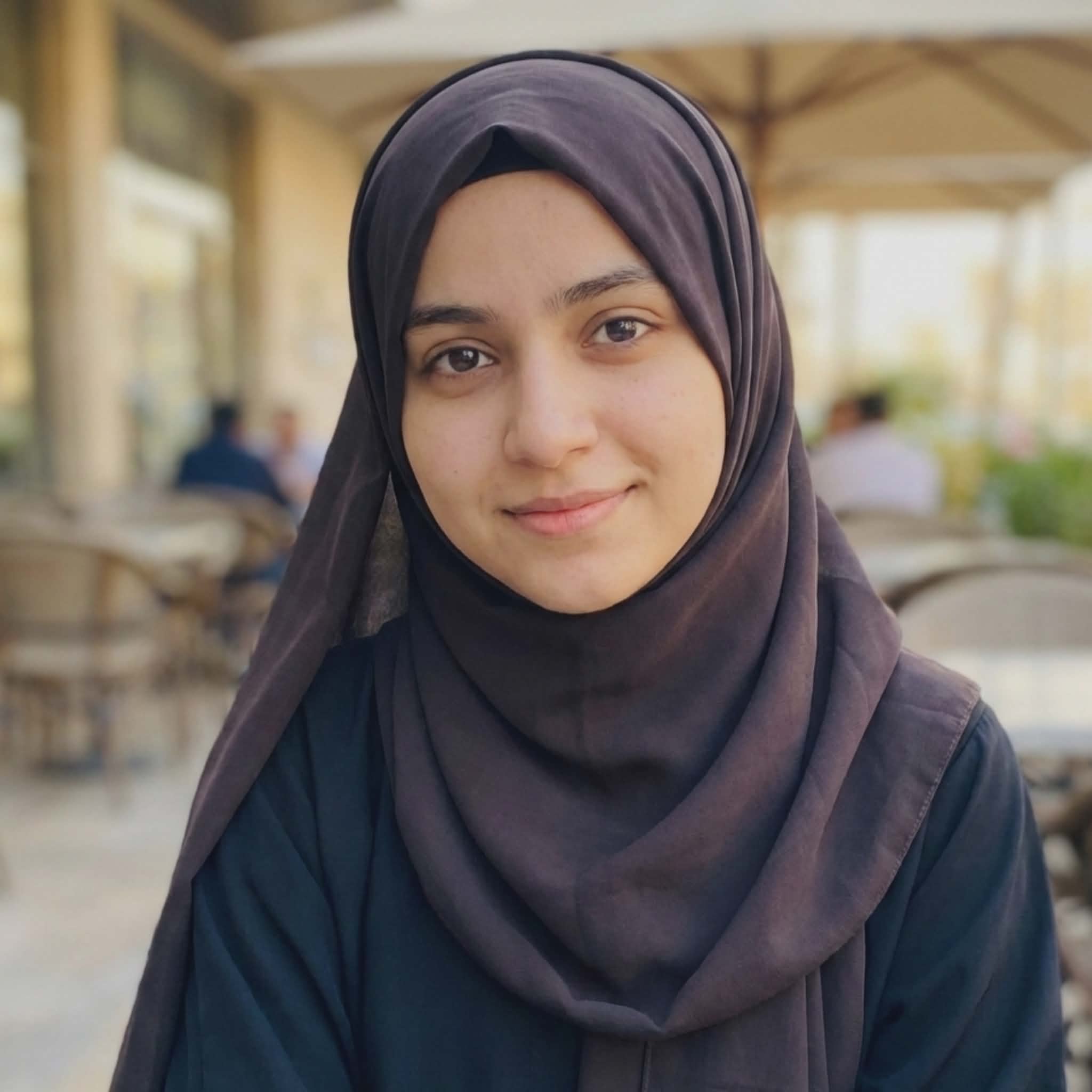 Portrait of a beautiful Muslim girl wearing a simple black hijab, smiling gently at the camera, blurred outdoor cafe background, modest fashion profile picture.